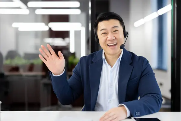 Portrait of happy asian male in business suit and headphones waving hello with hand while looking at camera. Excited man saying hi or goodbye during remote conversation with international partners.