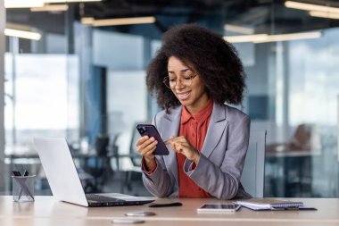 Modern ofis ortamında akıllı telefonunu kullanan odaklanmış bir Afro-Amerikan iş kadını. Çevresi teknoloji ile çevrili, kurumsal bir ortamda bağlantı ve verimliliği gösteriyor..