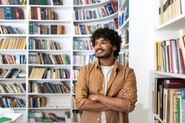 Neşeli bir gülümsemesi ve kıvırcık saçları olan genç bir adam, kitaplarla dolu parlak bir kütüphanede kollarını kavuşturup, bilgelik ve olumluluk hissi yayıyor..