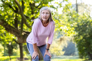 Yaşlı, kır saçlı, spor yapan, koşan, parkta yürüyen, elleri dizlerinin üstünde dinlenen, ağır nefes alan yaşlı bir kadın..