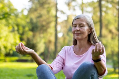 Park dışında yoga yapan, Lotus pozisyonunda oturan, gözleri kapalı meditasyon yapan yaşlı gri saçlı kadının yakın plan fotoğrafı..