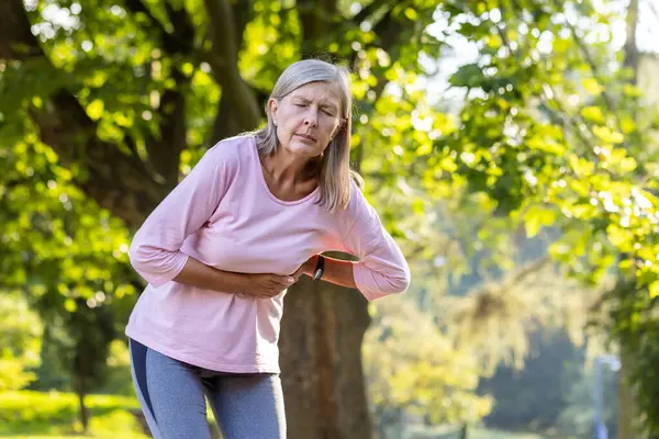 An older gray-haired woman is standing in the park bent over and holding her hands to the side of her body, feeling severe pain, needing medical help and rest.