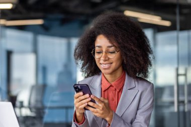 Neşeli bir Afrikalı genç iş kadını, teknoloji ve profesyonelliğin karışımını sergileyen çağdaş bir ofis ortamında akıllı telefonuyla meşgul oluyor..