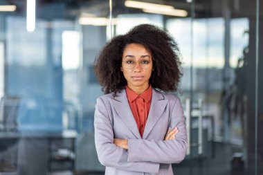 Ofiste gözlüklü ve takım elbiseli, kollarını göğsüne dolayıp kameraya ciddi ciddi bakan kendinden emin ve başarılı bir Afrikalı Amerikalı iş kadınının portresi..