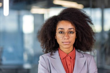 Ciddi ve başarılı, takım elbiseli ve gözlüklü Afro-Amerikan bir kadının yakın plan portresi ofiste kendini beğenmiş bir şekilde kameraya bakıyor..