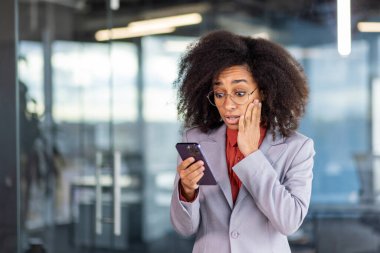 Şok olmuş genç Afrikalı Amerikalı kadın ofis yerinde duruyor ve telefon ekranına bakıyor, şaşkınlıkla, yüz yüze..