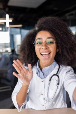 Bir klinikte genç bir Afro-Amerikalı kadın doktorun kameraya yaptığı video konuşmasında el sallayıp gülümserken dikey yakın plan fotoğrafı..
