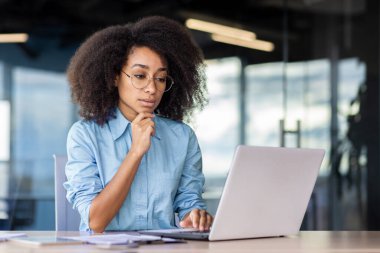 Ciddi bir Afro-Amerikalı kadın ofiste mavi gömlekli bir şekilde oturuyor ve özenle dizüstü bilgisayarına bakıyor..