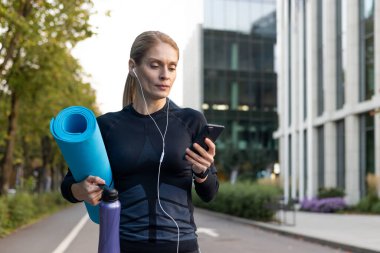 Yoga minderi ve hidrasyon taşıyan odaklanmış bir bayan atlet, müzik veya podcast dinliyor ve şehir ortamındaki bir spor salonunda açık hava fitness seansından önce akıllı telefonunu kontrol ediyor..