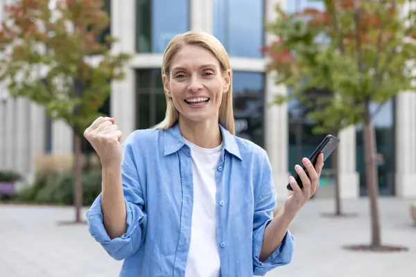 A joyous moment captured as a person celebrates success or good news outdoors, holding up a fist in a triumphant gesture with a smartphone in the other hand, depicting victory, achievement, and
