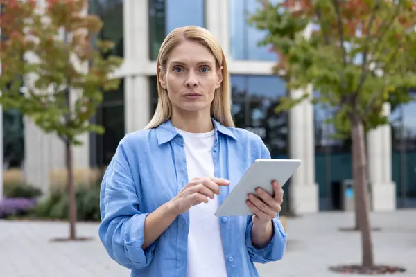 A focused professional woman stands outside on a cloudy day, using a digital tablet in front of a contemporary office building, embodying modern business and technology.