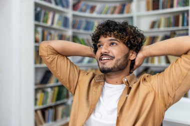 Genç bir insan, huzurlu bir kütüphane ortamında ellerini kitapların arasına koyup dinleniyor..