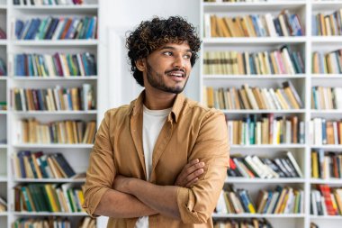 Sıradan giyinmiş bir profesyonel kesinlikle kitaplığın önünde kollarını kavuşturmuş duruyor. Konferansta eğitim veya edebiyat ortamında güven ve günlük profesyonellik üzerinde duruluyor.