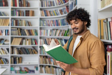 Yetişkin bir erkek, yeşil bir kitap okumaya dalmış, iyi aydınlatılmış bir kütüphanede duruyor. Kitaplıklar onu çevreleyen çeşitli kitaplarla dolu. Çalışma ve edebiyat araştırmaları için sakin bir ortam izlenimi uyandırıyor..