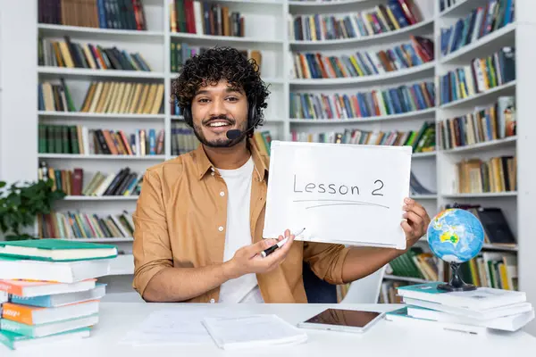 A young, engaging male teacher with a headset smiles as he presents Lesson 2 written on a whiteboard, surrounded by stacks of books in a bright library environment, symbolizing education