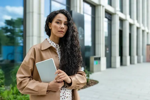 A confident, curly-haired professional woman stands outside a contemporary office building, holding a tablet and looking pensive. She appears focused and ready for business in a stylish outfit.