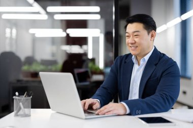 Başarılı ve gülümseyen Hintli genç adam modern bir ofiste takım elbise içinde oturuyor ve dizüstü bilgisayarda çalışıyor, klavyede mesaj yazıyor ve daktilo ediyor..