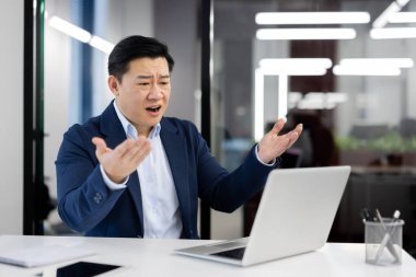 Hayal kırıklığına uğramış takım elbiseli Asyalı bir adam dizüstü bilgisayarın önünde oturuyor ve kolları açık bir şekilde ekrana bakıyor..