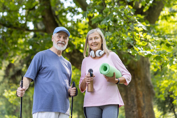 Portrait of a scary couple, gray-collared man and woman doing sports in the park, standing with trekking poles, holding a mat and a bottle of water. They look and smile at the camera.