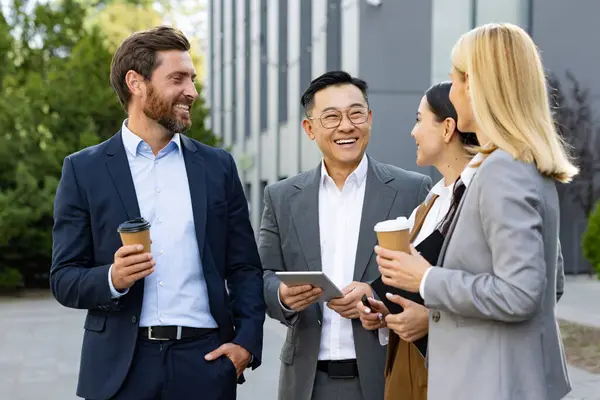 Four cheerful colleagues in business attire with takeaway coffee, engaging in a friendly conversation outside a modern office.