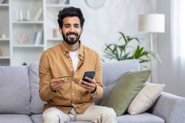 İyimser çeşitlilikteki erkek koltukta oturuyor ve elinde cep telefonu ve kredi kartıyla kameraya gülümsüyor. Bankacılık başvurusuyla anında para transferi yapan mutlu sakallı adam..