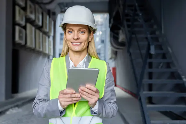 Professional caucasian woman in helmet and neon yellow vest standing at construction with digital tablet in hands. Experienced female architect checking house building process and entering data.