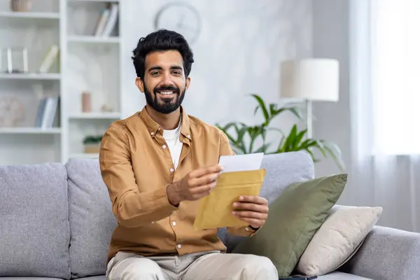 Smiling latino man receiving postal letter and opening it at home while sitting on grey pillowed sofa. Brunette male getting mail with greeting card and formal invitation for long waited event.