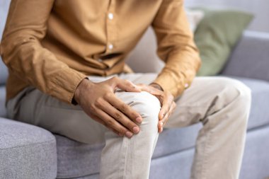 Gri kanepede minderlerle otururken iki elinle dizini tutan tanımadığın kişiye yaklaş. Bacak travması olan yetişkin bir adam eklem ağrısı çekiyor ve masaj için hassas bir nokta..