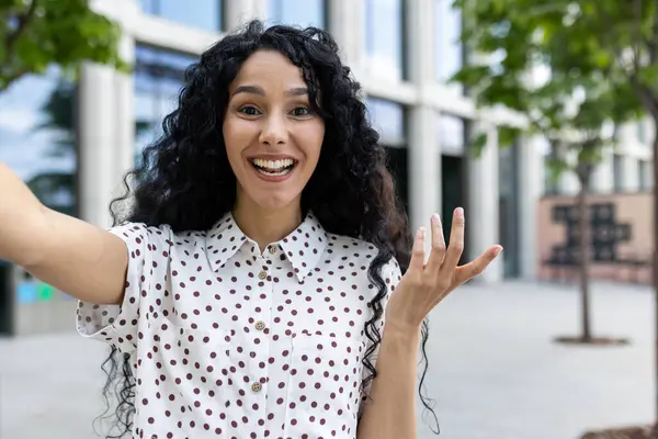 Happy curly woman in polka dot blouse recording video and holding camera in spreaded hand while walking in city centre. Satisfied female filming vlog about new shared coworking for freelancers.