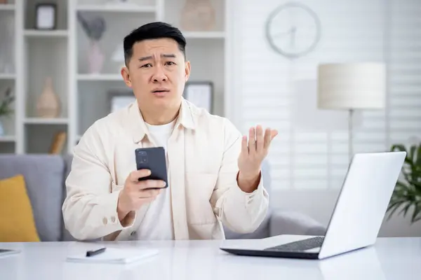Disturbed chinese male in casual clothes holding mobile phone while working at home office with portable computer. Unhappy remote manager receiving text message with penalty in corporate chat.