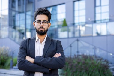 Kollarını kavuşturmuş iyi giyimli bir iş adamı, çağdaş bir ofis ortamının dışında inançla ayakta duruyor. Güven ve profesyonellik yayarak çeşitli kurumsal başarıları temsil ediyor..