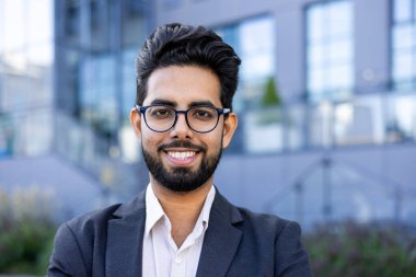 Sivri takım elbiseli genç bir Hintli işadamı arka planda kentsel bir manzara ile güvenli bir şekilde dışarıda duruyor. Kurumsal çeşitlilik temaları için ideal.