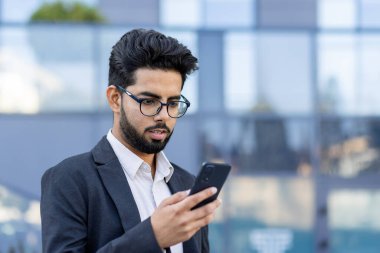 Takım elbiseli, akıllı telefonuna odaklanmış genç bir profesyonel dışarıda duruyor. Bu görüntü şehir ortamında modern iş iletişimini büyülüyor. Bağlantıyı tasvir etmek için mükemmel.