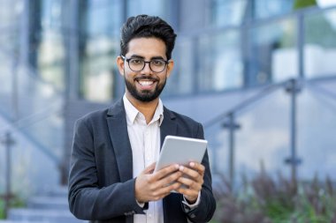 Akıllı takım elbiseli genç bir işadamı tablet bilgisayarını kullanmaya dalmış. Çağdaş bir ofis ortamının dışında, küresel iş ve bağlantıyı sembolize ediyor..