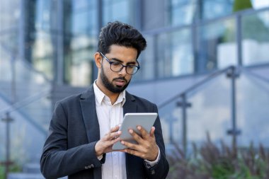 Gözlük takan Hintli profesyonel bir işadamı modern cam binaların dışında tablet kullanıyor. Küresel bir ortamda mobil iş iletişimi kavramı resmedilmiştir.