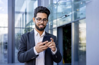 Takım elbiseli genç bir Hintli işadamı akıllı telefonuyla meşgul, mesaj atarken ya da mesaj okurken gülümsüyor. Bu görüntü modern profesyonel hayatın bir anını yakalar, muhtemelen bir mola sırasında ya da...