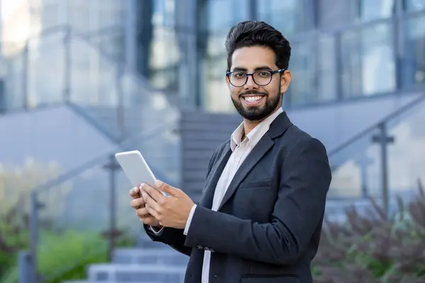A cheerful Indian businessman in formal attire stands outdoors, using a digital tablet with a confident smile. Modern business setting with a multicultural subject.