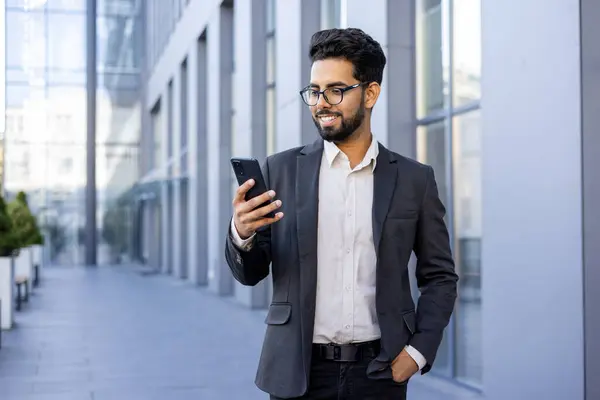 A happy and successful young Indian man in formal wear is engaged with his smartphone, smiling while standing in a modern city environment. This image captures the essence of connectivity, urban life