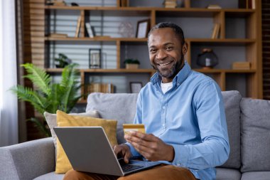 Neşeli Afro-Amerikan erkek e-ticaret için dizüstü bilgisayar kullanıyor, kredi kartı tutuyor. Ev konseptinde online alışveriş.
