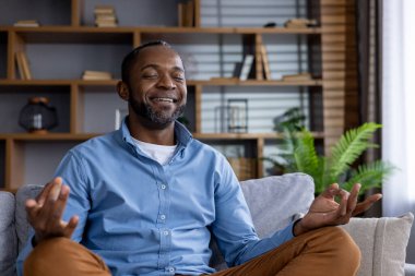 Afro-Amerikalı bir adam günlük giysiler içinde gözleri kapalı oturuyor, gülümsüyor ve huzurlu bir oturma odasında meditasyon yapıyor..