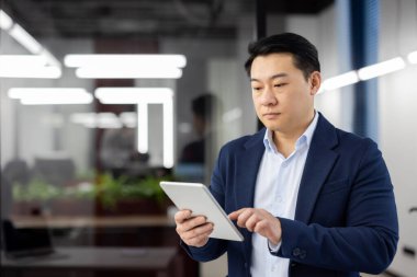 Modern bir ofis alanında odaklanmış Asyalı işadamı, günlük işinin bir parçası olarak dijital tablet kullanıyor..