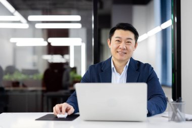 Mavi takım elbiseli Asyalı bir işadamı elinde dizüstü bilgisayarla oturmuş, parlak bir ofis ortamında güven ve profesyonellik yayıyor..