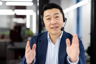 Takım elbiseli Asyalı bir işadamı modern bir iş yerinde konferans görüşmesi için kulaklık kullanıyor. Olumlu ifade ediyor..