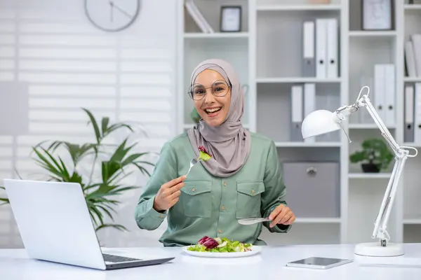 Stock image A joyful Muslim woman wearing a hijab eats a fresh salad at her desk, taking a break from work with her laptop and lamp beside.