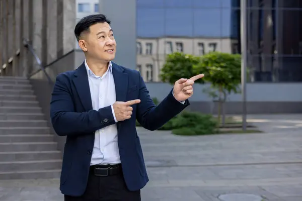 Asian businessman in suit pointing with both hands to side on a sunny day, standing outside with buildings in background.