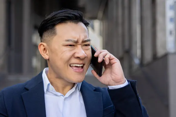 Annoyed Asian adult male in a business suit on a call outdoors, showing signs of frustration and urgency.