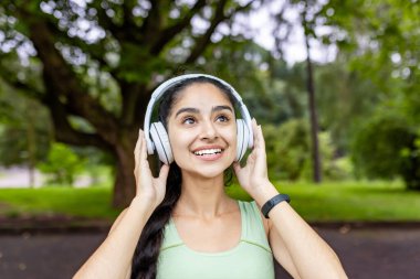 Gülümseyen, kulaklık takan ve eşofman giyen genç bir Hintli kadın müzikten zevk alıyor, açık hava fitness aktiviteleri için motive oluyor..