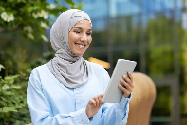 Professional happy Muslim woman with hijab holding a tablet, enjoying technology outside a modern office setting.