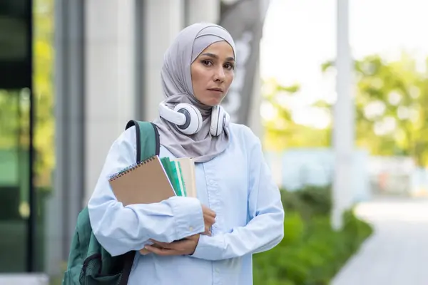 Portrait of a focused Muslim woman with hijab, headphones, and textbooks stepping outside in an academic setting.