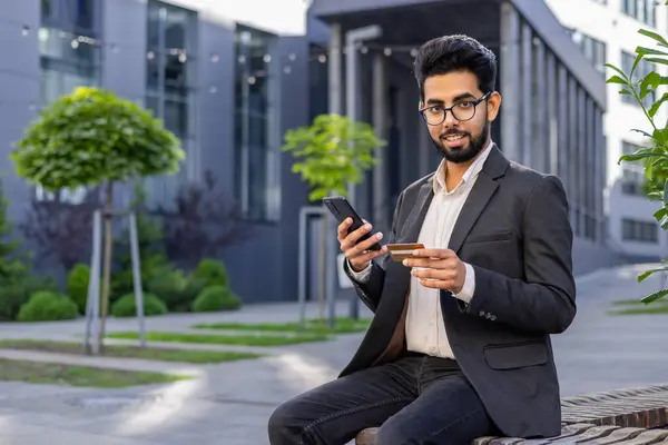 Portrait of a young Indian man in a business suit sitting on a bench near an office center, holding a credit card and a phone, looking at the camera with a smile.